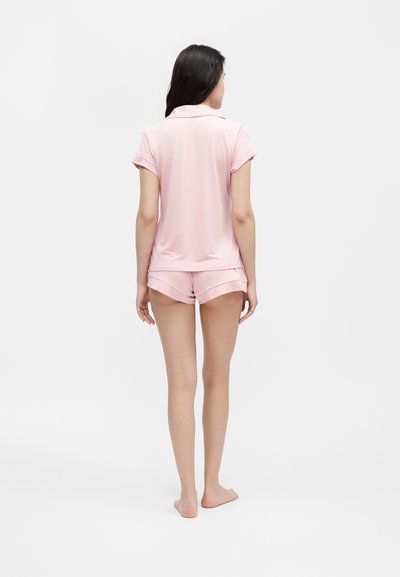 Γυναίκα όρθια ξυπόλητη με πλάτη, φοράει ροζ ανοιχτό κοντομάνικο τοπ και αντίστοιχα σορτς πιτζάμες σε απλό φωτεινό φόντο.