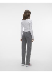 Femme debout de dos, portant un haut blanc à manches longues, un jean gris taille haute et des baskets blanches avec des détails rouges et bleus.