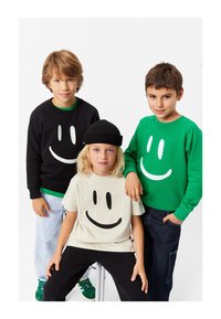 Tre bambini indossano abbigliamento informale con motivi di faccine sorridenti: una felpa nera, una felpa verde e una maglietta beige, tutte in tessuto morbido.