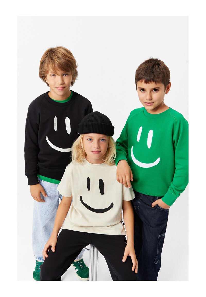 Tre bambini indossano abbigliamento informale con motivi di faccine sorridenti: una felpa nera, una felpa verde e una maglietta beige, tutte in tessuto morbido.