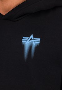 Svart hettegenser med et blått grafisk design som viser en kantete stjerne og to horisontale striper, som skaper en falmende effekt nederst.