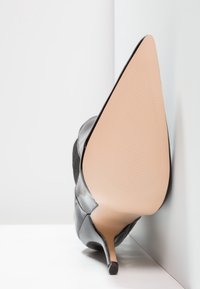 Scarpa nera con tacco alto, punta affusolata, superiore in pelle lucida verniciata e tacco a stiletto elegante. Suola in gomma nuda liscia con texture.