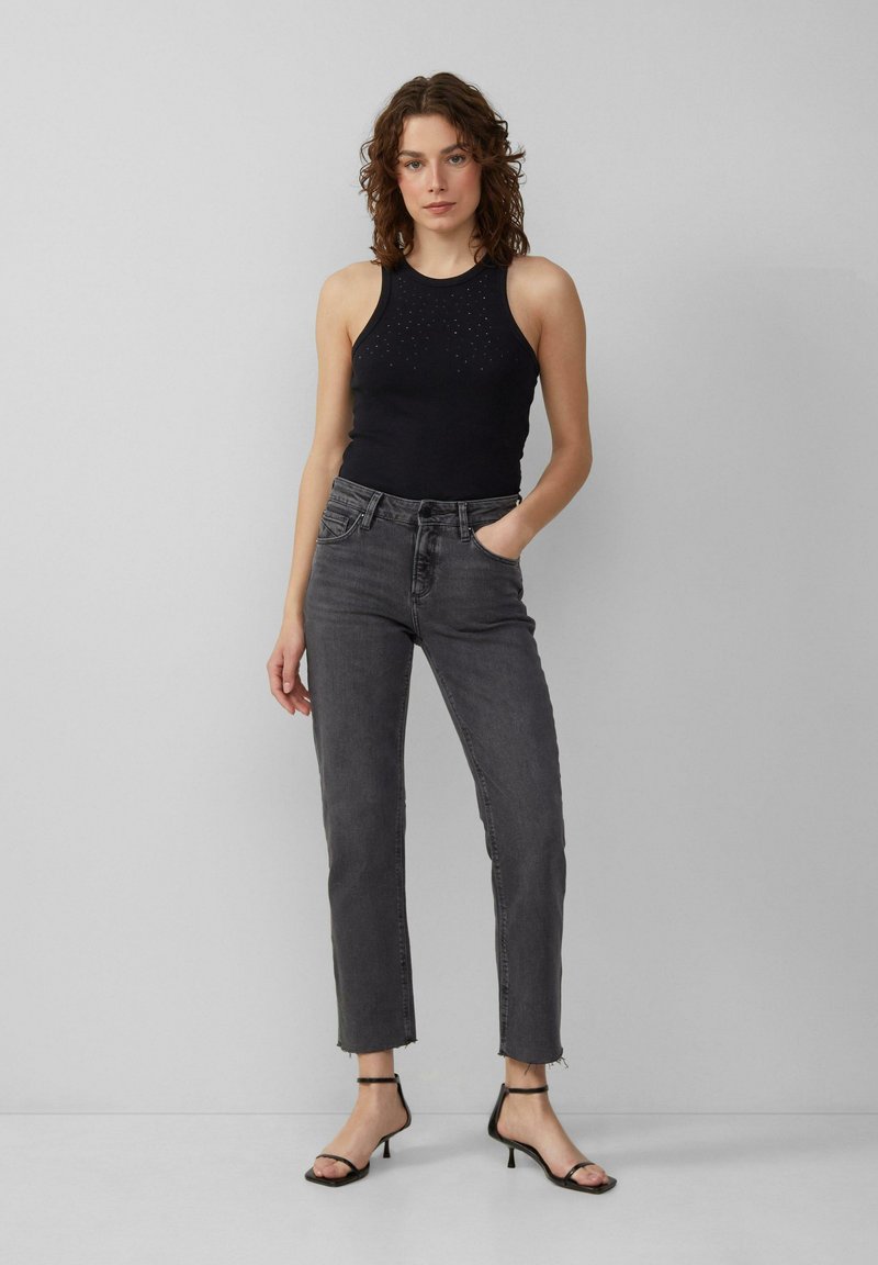 Haut sans manches noir avec un motif texturé, associé à un jean gris taille haute et des sandales à talons noires. Fond uni.