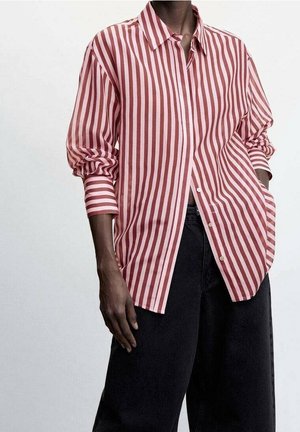 Personne portant une chemise ample à manches longues à rayures verticales rose et blanches, avec un pantalon noir à jambes larges, les mains détendues le long du corps.