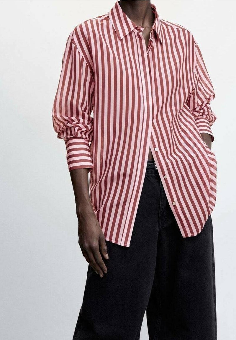 Personne portant une chemise ample à manches longues à rayures verticales rose et blanches, avec un pantalon noir à jambes larges, les mains détendues le long du corps.