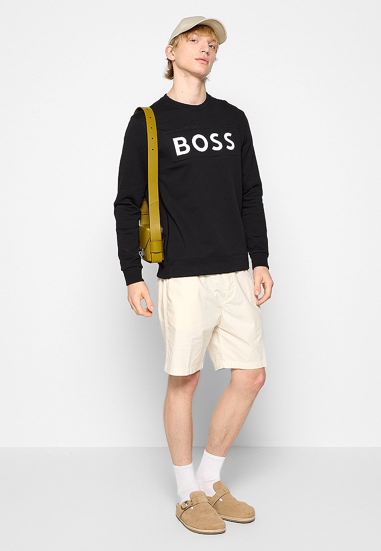 Felpa nera con logo "BOSS", pantaloni corti beige, scarpe slip-on color sabbia, calzini bianchi e un cappellino chiaro. Borsa a tracolla marrone.