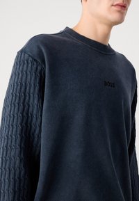 Granatowy sweatshirt z teksturowanymi rękawami, okrągłym dekoltem i subtelnym logo BOSS na piersi. Miękka tkanina z luźnym krojem.