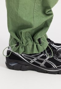 Crne atletske tenisice imaju teksturirani mrežasti gornji dio sa srebrnim naglascima. Zelene hlače imaju suženi donji dio s detaljem užeta i rebrastom teksturom.