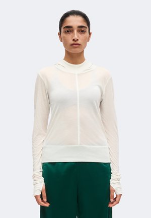 Femme aux cheveux foncés attachés, portant un haut blanc transparent à manches longues et un pantalon vert foncé, debout sur un fond uni.