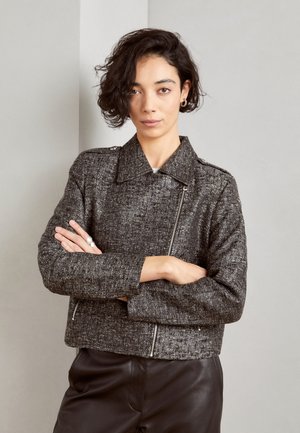 Giovane donna con capelli ricci e corti, che indossa una giacca grigia con zip e pantaloni neri, in piedi con le braccia incrociate su uno sfondo semplice.