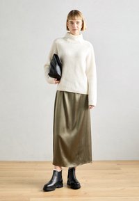 Pull à col roulé côtelé crème, jupe maxi en satin olive, et bottines en cuir noir. Le modèle tient un sac noir, debout sur un sol en bois.