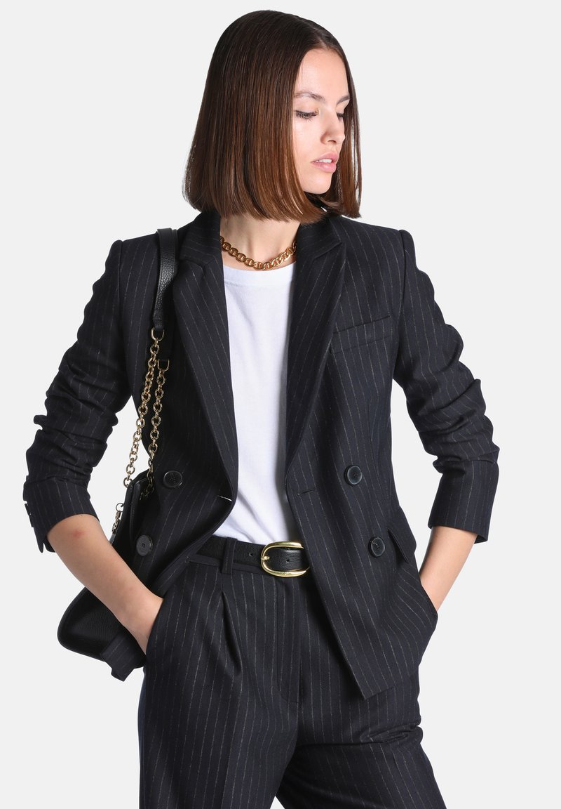 Femme aux cheveux bruns mi-longs porte un blazer et un pantalon noirs à fines rayures, une chemise blanche, un collier chaîne doré et un sac noir avec une bandoulière chaîne dorée.