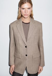 Beige op maat gemaakte blazer met een gestructureerde stof, twee zakken aan de voorkant en zwarte knopen. Draagt over een bruine en witte gestreepte blouse.