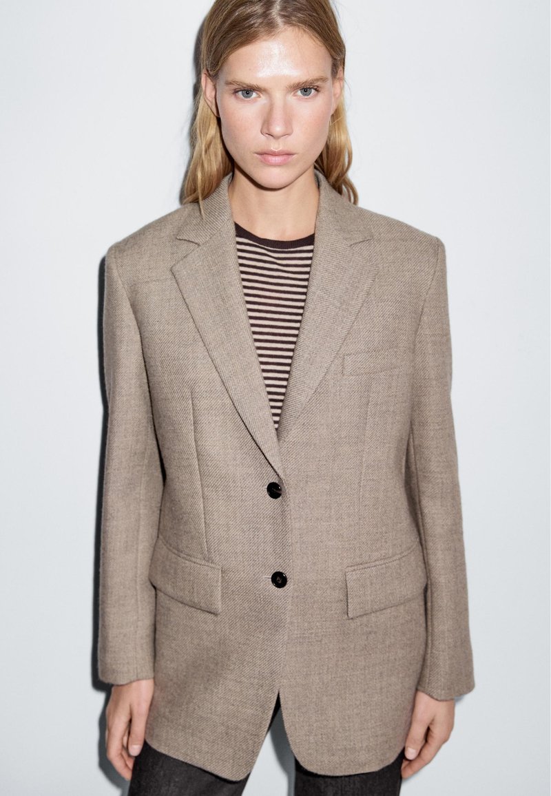 Beige op maat gemaakte blazer met een gestructureerde stof, twee zakken aan de voorkant en zwarte knopen. Draagt over een bruine en witte gestreepte blouse.