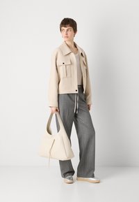 Veste courte beige avec de grandes poches, associée à un pantalon large rayé gris et des chaussures argentées à enfiler. Porte un sac hobo beige.