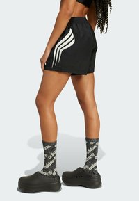 Fekete rövidnadrág fehér csíkokkal, rugalmas derékpánttal, fekete textúrált klumpákkal és sötét mintás zoknikkal, amelyek logót viselnek.