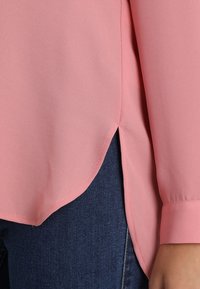 Blouse rose en tissu léger, avec un ourlet arrondi, des manches longues et des fentes latérales. Associée à des matériaux en denim foncé.