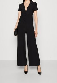 Svart jumpsuit med korta ärmar, djup V-ringning och vida ben. Tillverkad av ett mjukt tyg, med en framåtvänd vridning som detalj.