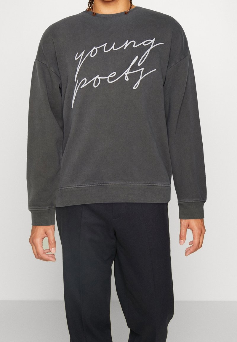 Personne portant un sweat-shirt gris foncé avec "young poetry" écrit en cursive et un pantalon noir, debout devant un fond uni.