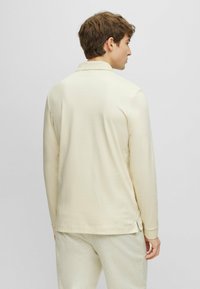 Hosszú ujjú, galléros ing világos krémszínben. Simább textúra, slim fit szabás, oldalsó résnyílások az alján, tűzött mandzsetták, könnyű anyag. Diszkrét fényes felület.