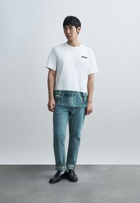 Άνδρας που φοράει λευκό T-shirt της Moschino με εκτύπωση σχεδίου τζιν στο κάτω μέρος, μπλε τζιν, γκρι κάλτσες και μαύρα μοκασίνια, όρθιος.