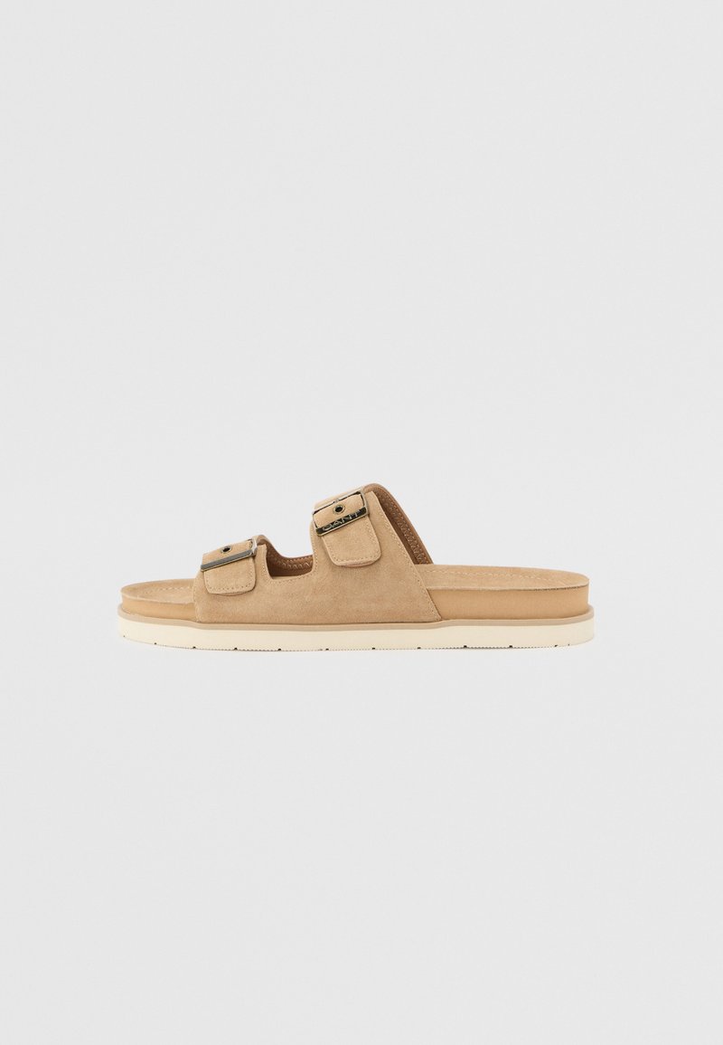 Sandales à glisser en suède beige avec deux sangles réglables dotées de boucles métalliques, une semelle intérieure rembourrée et une semelle légère blanche.