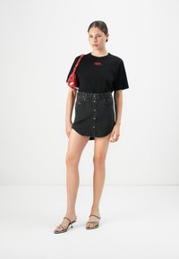 Kvinne i svart t-skjorte med logo, svart denimskjørt med knapper foran, sandaler med stropper og hæler, og bærer en liten rød skulderveske.