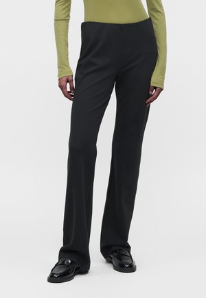 Persona che indossa pantaloni neri a zampa, mocassini neri lucidi e una maglia aderente a maniche lunghe verde oliva, in piedi davanti a uno sfondo bianco semplice.