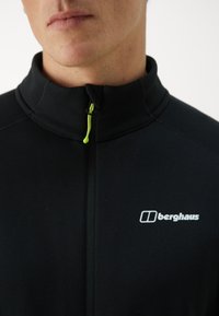 Giacca nera con zip alta, dotata di un tirazip verde e un logo Berghaus bianco sul lato sinistro. Tessuto morbido.