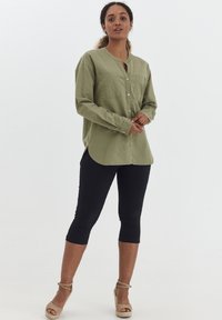 Camisa de botón verde oliva con cuello, dos bolsillos en el pecho y dobladillo curvado, combinada con leggings capri negros y sandalias de cuña beige.