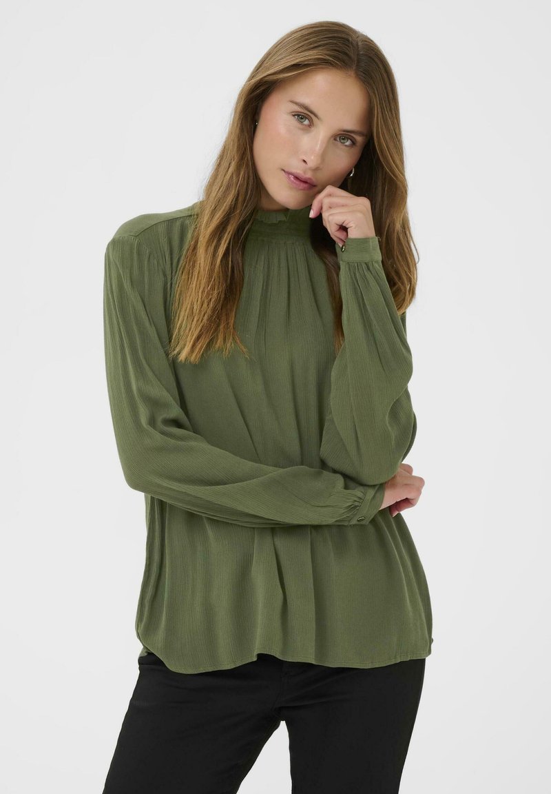 Bluză lungă, cu mânecă lungă, de culoare verde măslin, cu guler înalt, textură pliată, manșete cu buton și croială lejeră, asortată cu pantaloni negri.