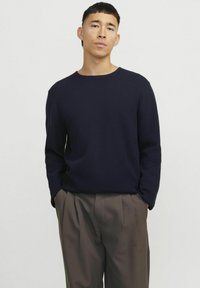 Suéter de punto azul marino con cuello redondo y mangas largas, combinado con pantalones plisados de un marrón claro. Textura suave, corte ajustado.