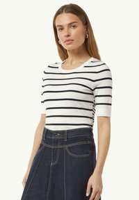 Gestreiftes, kurzärmliges Strickoberteil in Weiß und Marineblau; mit rundem Ausschnitt und taillierter Silhouette. Getragen mit einem Jeansrock.