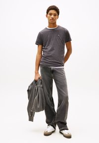 Šedé pletené tričko s bílým kulatým výstřihem, v kombinaci se šedými džínami z denimu. Model drží šedou mikinu s kapucí a má na sobě bílé a hnědé tenisky.