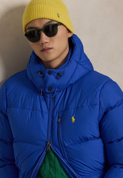 Modra puhasta jakna s kapuco z rumeno nalepko, zadrgo spredaj in nastavljivimi vrvico. Oblečena s zelenim puloverjem in rumeno kapa.