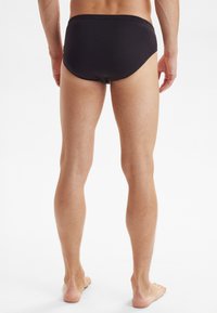 Costumi da bagno neri con vita elastica, design aderente e texture liscia, mostrati da dietro, con gambe e piedi nudi.