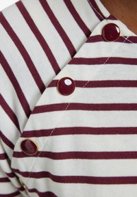 Gestreiftes Oberteil in bordeauxrot und weiß, mit diagonellem Ausschnitt und großen bordeauxroten Knöpfen an der Schulter. Weiche, strukturierte Stoffqualität.