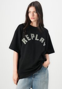 Svart bomull T-skjorte med korte ermer og avslappet passform. Har en iøynefallende "REPLAY" logo i grått over brystet. Kombinert med blå denimjeans.