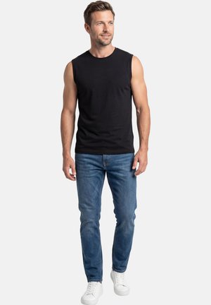Man standing wearing a black sleeveless shirt, blue jeans, and white sneakers, looking slightly to the side against a plain background.