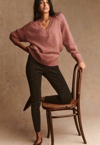 Žena u dusty rose V-izrez puloveru i crnim uskim hlačama, koljena na drvenoj stolici, naslonjena na jednostavnu bež pozadinu.