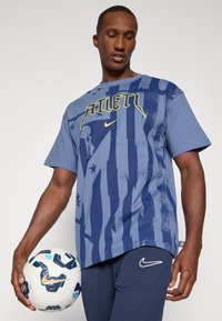 Blå bomulds T-shirt med et gult "ATLET" grafik på forsiden, med et stribet design og subtile mønstre, båret af en model der holder en fodbold.