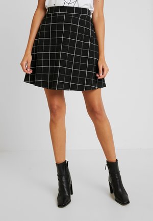 Jupe noire à motif quadrillé blanc, coupe évasée, longueur genou, fabriquée dans un tissu texturé. Associée à des bottines à talons noires.