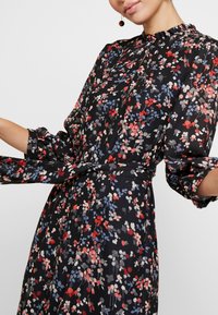 Robe florale avec un fond noir, ornée de motifs floraux rouges, roses et bleus. Elle a un col à volants, des manches longues et une ceinture en tissu.