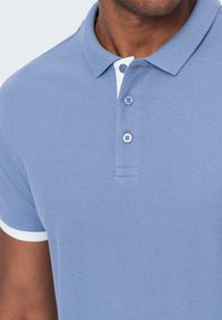 Svetle modrá polo košile s krátkým rukávem, s klasickým límcem a tří knoflíkovou légou. Bílé akcenty na límci a rukávech.