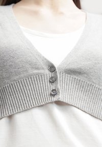 Světle šedý pletený cardigan s ribovým vzorem, se širokým výstřihem do V a čtyřmi tmavě šedými knoflíky. Nosit přes bílou košili.