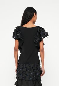 Musta paita, jossa on teksturoidut, röyhelöiset hihat ja kukkakoristeet. Sileä kangas, rento istuvuus, korkea kaulus ja avoin selkä.