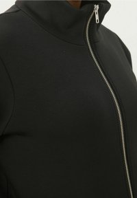 Veste noire à fermeture éclair avec col montant, texture lisse, coupe ajustée et zip argenté en avant.