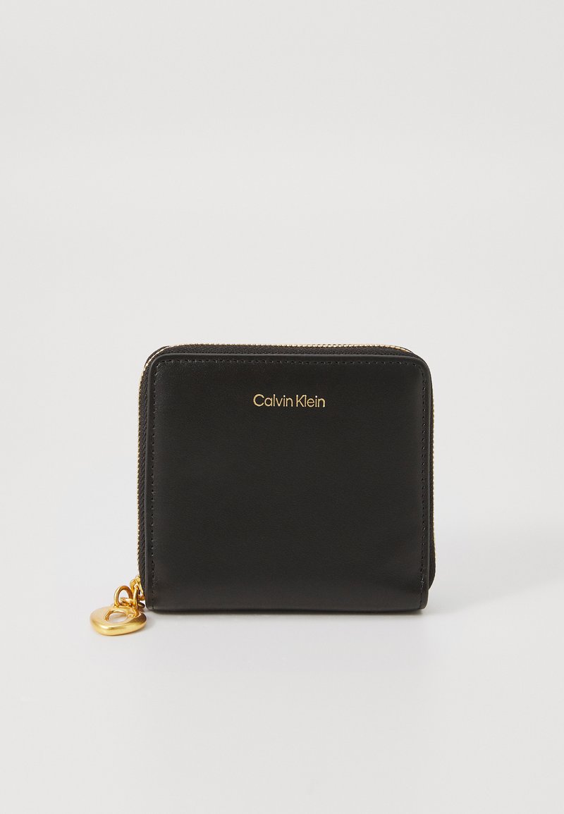 Portefeuille en cuir noir avec une fermeture éclair dorée et un logo, de forme rectangulaire, texture lisse, avec un accent en métal en forme de cœur.