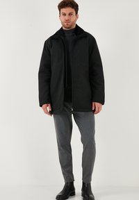 Crna flis jakna s visokim ovratnikom, zatvorena patentnim zatvaračem, s dva bočna džepa, u paru s sivim slim-fit hlačama i crnim gležnjačama.