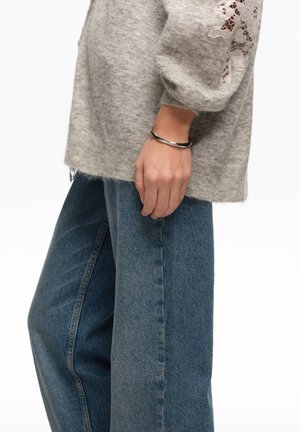 Persona con vaqueros azules holgados, un suéter gris con detalle de encaje en la manga y una pulsera plateada torcida en la muñeca izquierda.